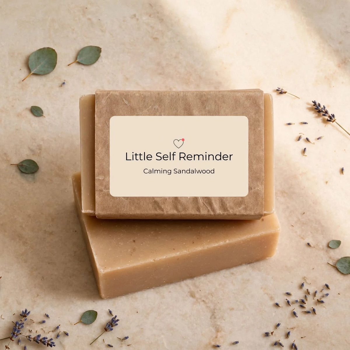 Two rectangular bars of handmade soap stacked on a light beige stone surface, with a cream label reading 'Little Self Reminder' and 'Calming Sandalwood'.