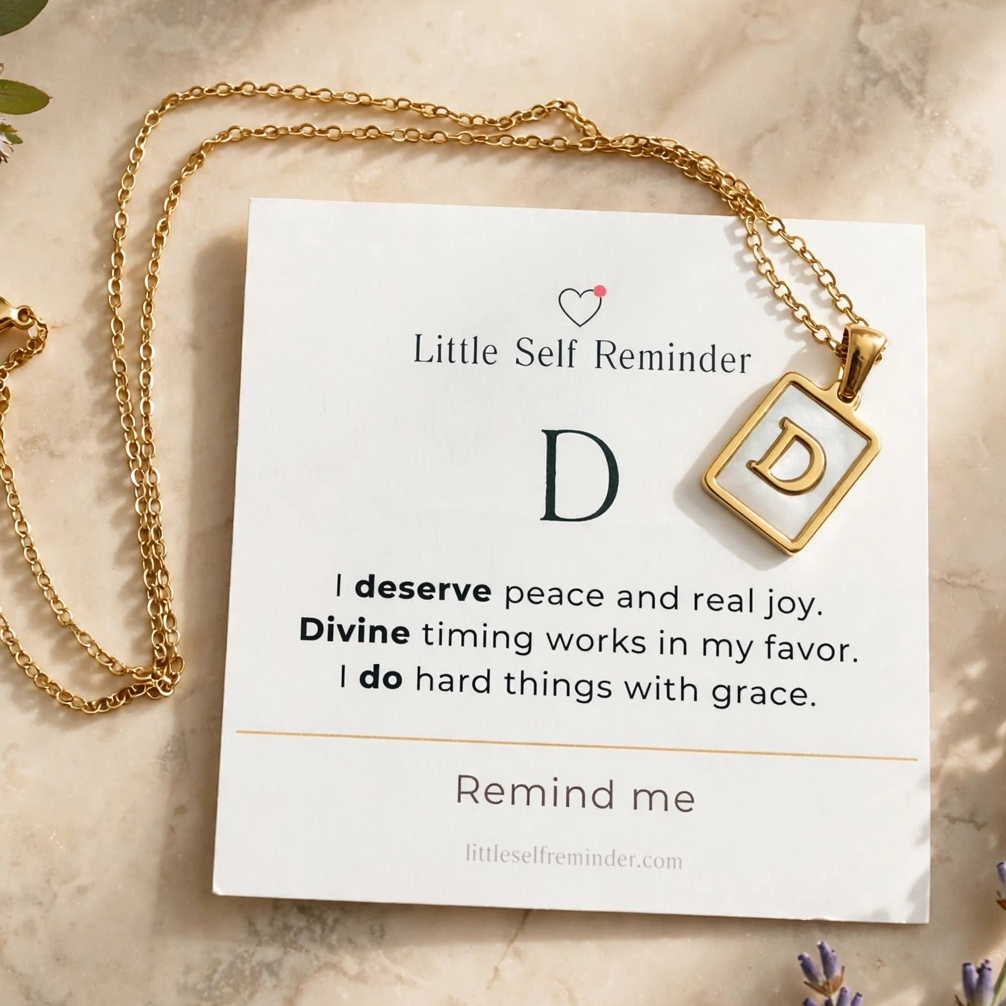 A gold necklace with a square pendant featuring the letter 'D' is displayed on a white card that reads 'Little Self Reminder' with a quote.
