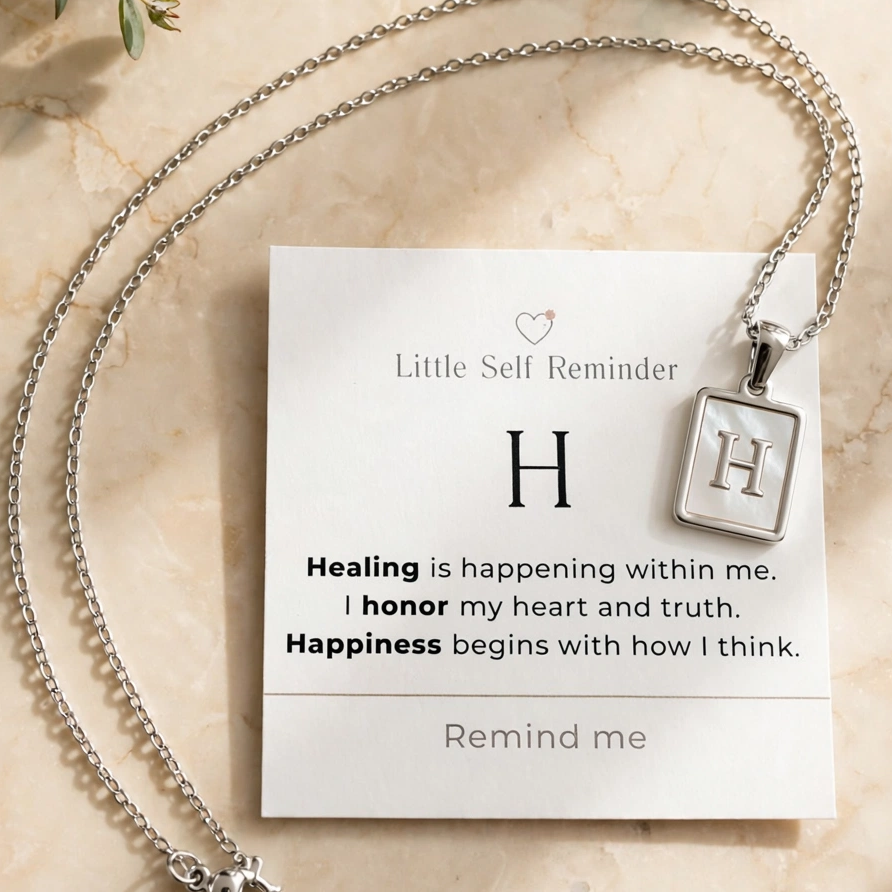 A silver initial necklace with a square pendant displaying the letter 'H', presented on a white 'Little Self Reminder' card with a quote.
