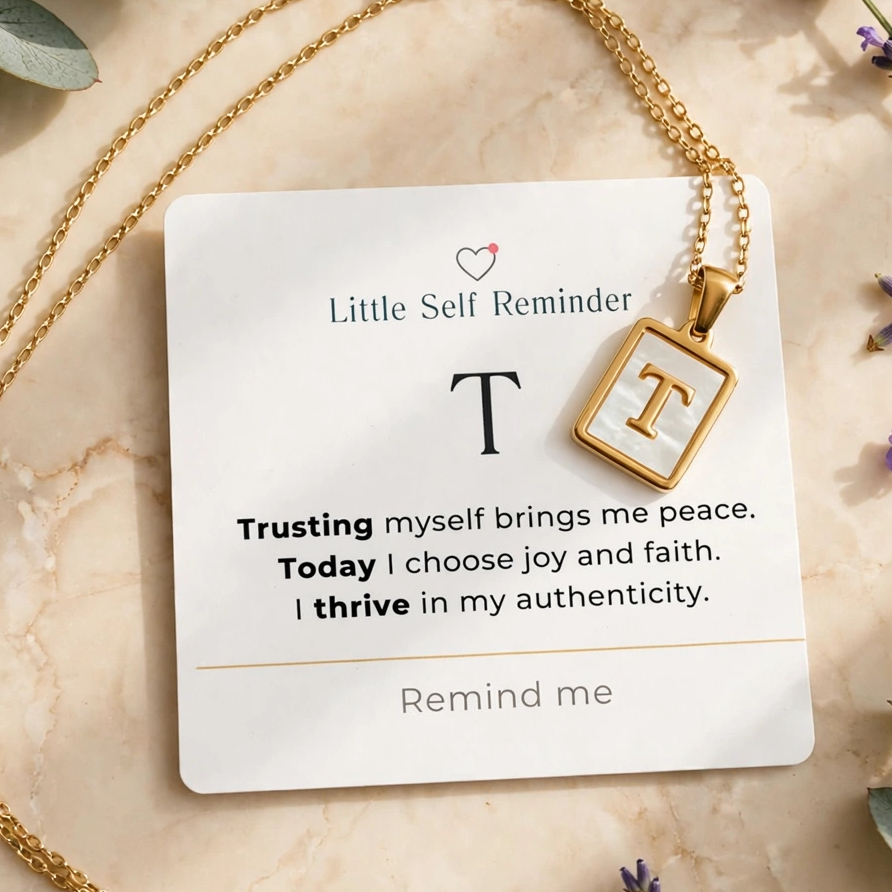 A gold necklace with a square pendant featuring the letter 'T' is displayed on a white card that reads 'Little Self Reminder' with a positive affirmation.