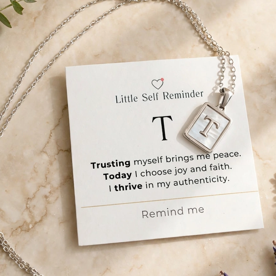 A silver necklace with a square pendant displaying the letter 'T' is draped over a white card that reads 'Little Self Reminder'.
