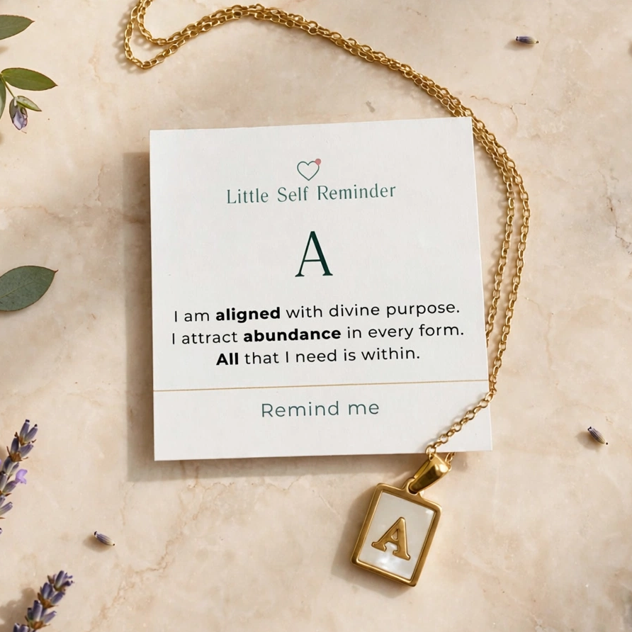 A gold initial necklace with a square pendant holding the letter 'A' is displayed on a cream card that reads 'Little Self Reminder' with a quote starting with 'A'. 