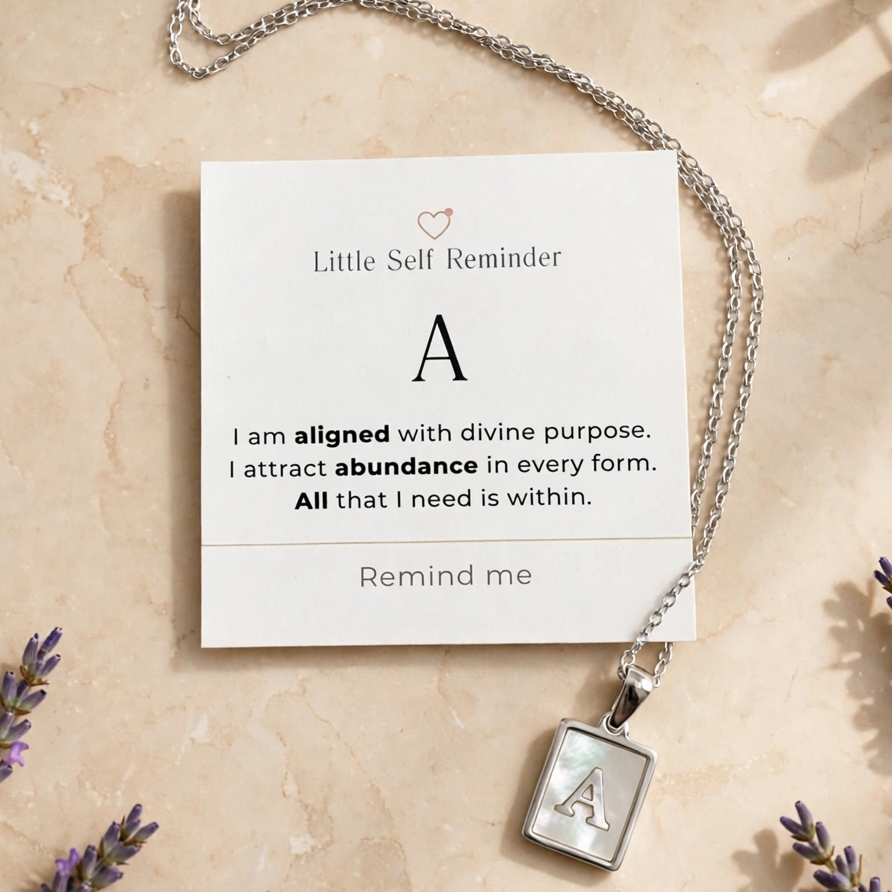 A silver necklace with a square pendant engraved with the letter 'A' is displayed on a cream card that reads 'Little Self Reminder' with a supportive affirmation.