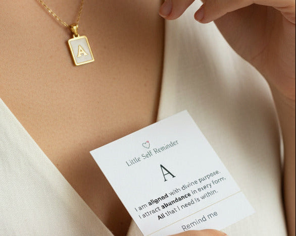 A close-up lifestyle shot of a model wearing pendant necklace with the letter 'A' on it, while holding a white card that reads 'Little Self Reminders'.