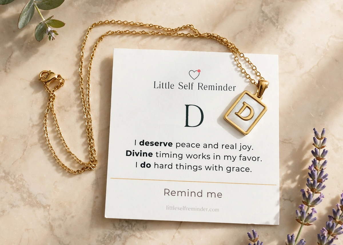 A gold necklace with a square pendant featuring the letter 'D' is displayed on a white card that reads 'Little Self Reminder' with a quote.