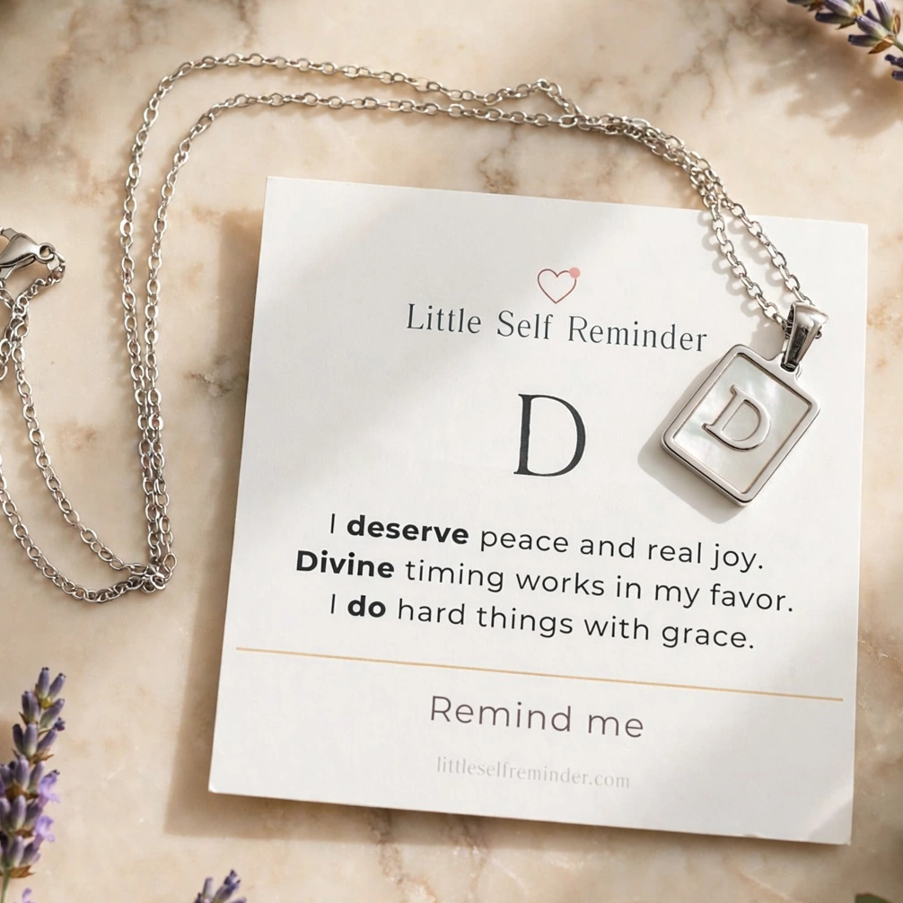 Silver necklace with a square pendant on a white card that reads 'Little Self Reminder' with a letter D and a quote.