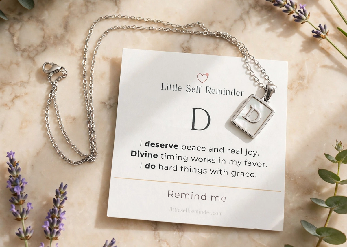 Silver necklace with a square pendant on a white card that reads 'Little Self Reminder' with a letter D and a quote.
