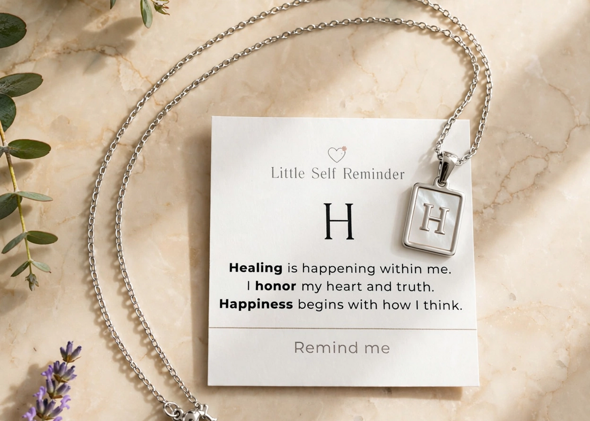 A silver initial necklace with a square pendant displaying the letter 'H', presented on a white 'Little Self Reminder' card with a quote.