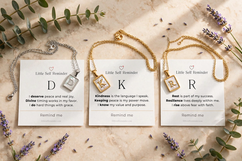 Three gold and silver necklaces with square pendant cards are arranged on a beige fabric background with eucalyptus leaves and lavender sprigs.