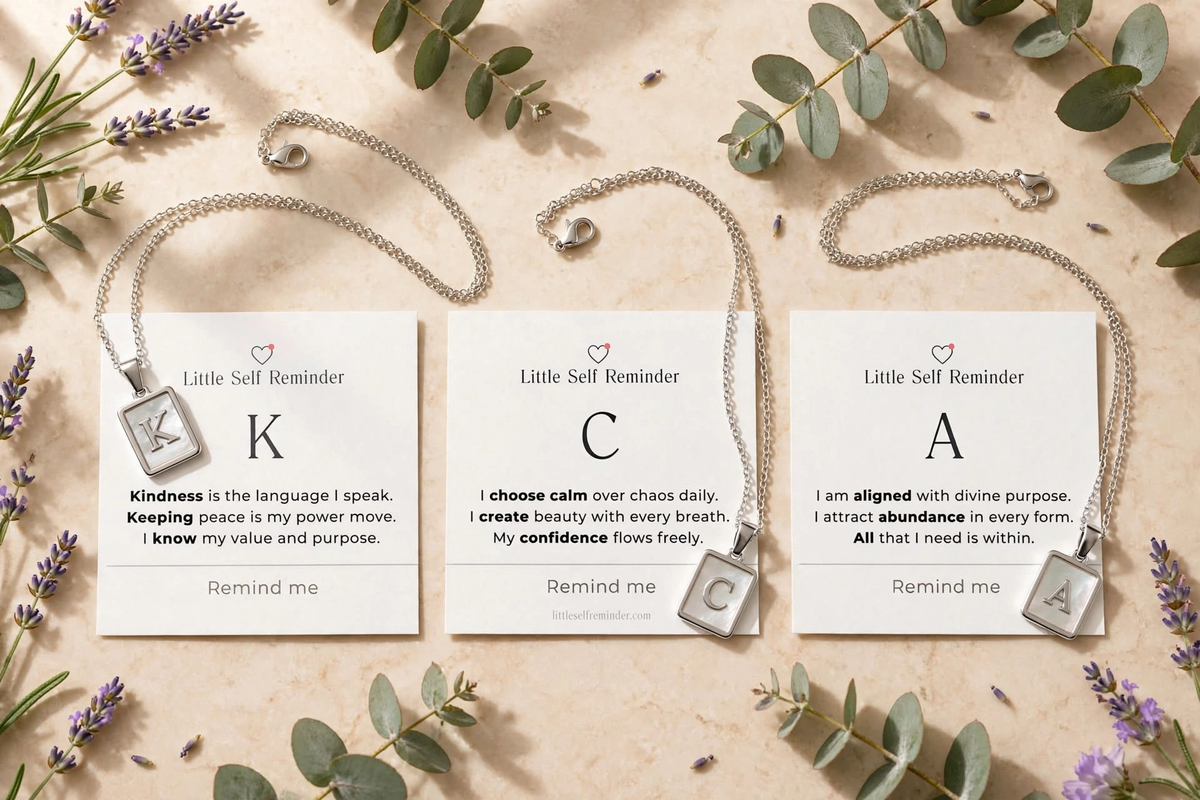 Three silver necklace pendants with square letter charms are arranged on a beige surface with eucalyptus leaves and lavender sprigs.