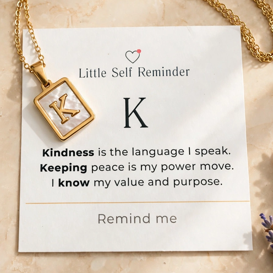 A gold necklace with a square pendant featuring a large letter K and a small heart icon, displayed on a cream card that reads 'Little Self Reminder'.