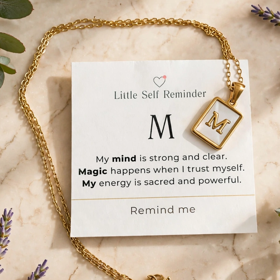 A gold necklace with a square pendant engraved with the letter 'M' is displayed on a white card that reads 'Little Self Reminder' with a motivational quote. 