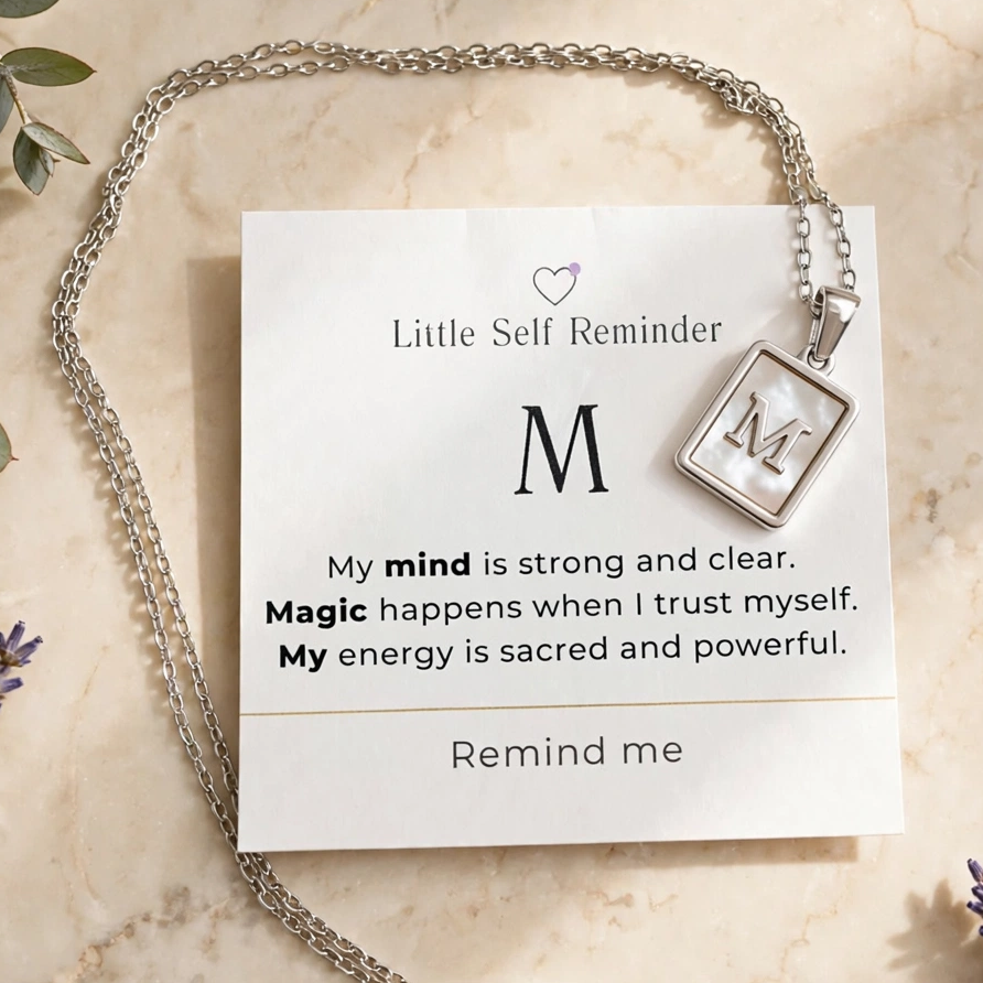 A silver chain necklace with a square pendant displaying the word 'REMIND' is arranged on a cream marble surface with dried flowers and greenery.