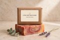 A Kraft paper box labeled 'Little Self Reminder' and 'Sweet Skin Berry' sits on top of a rectangular bar of soap with pink, orange, and cream swirls. 