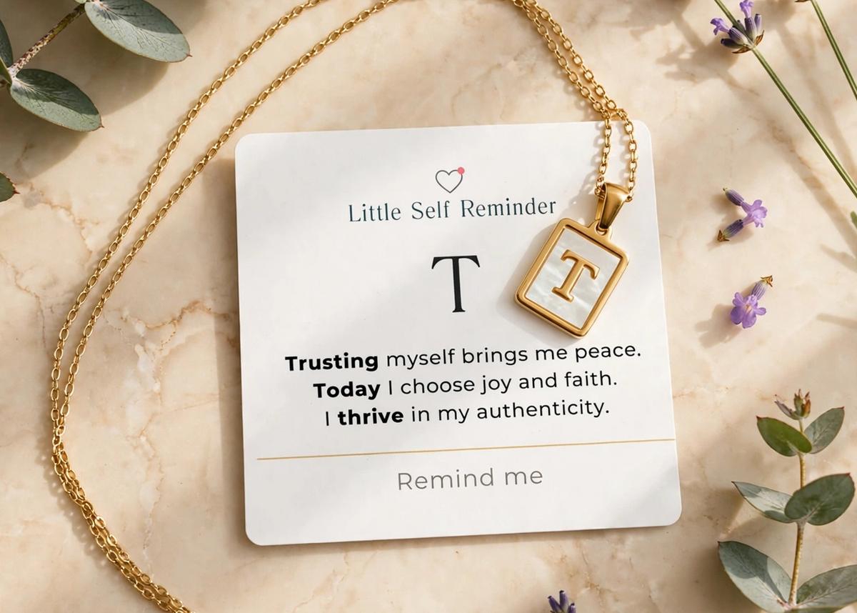 A gold necklace with a square pendant featuring the letter 'T' is displayed on a white card that reads 'Little Self Reminder' with a positive affirmation.