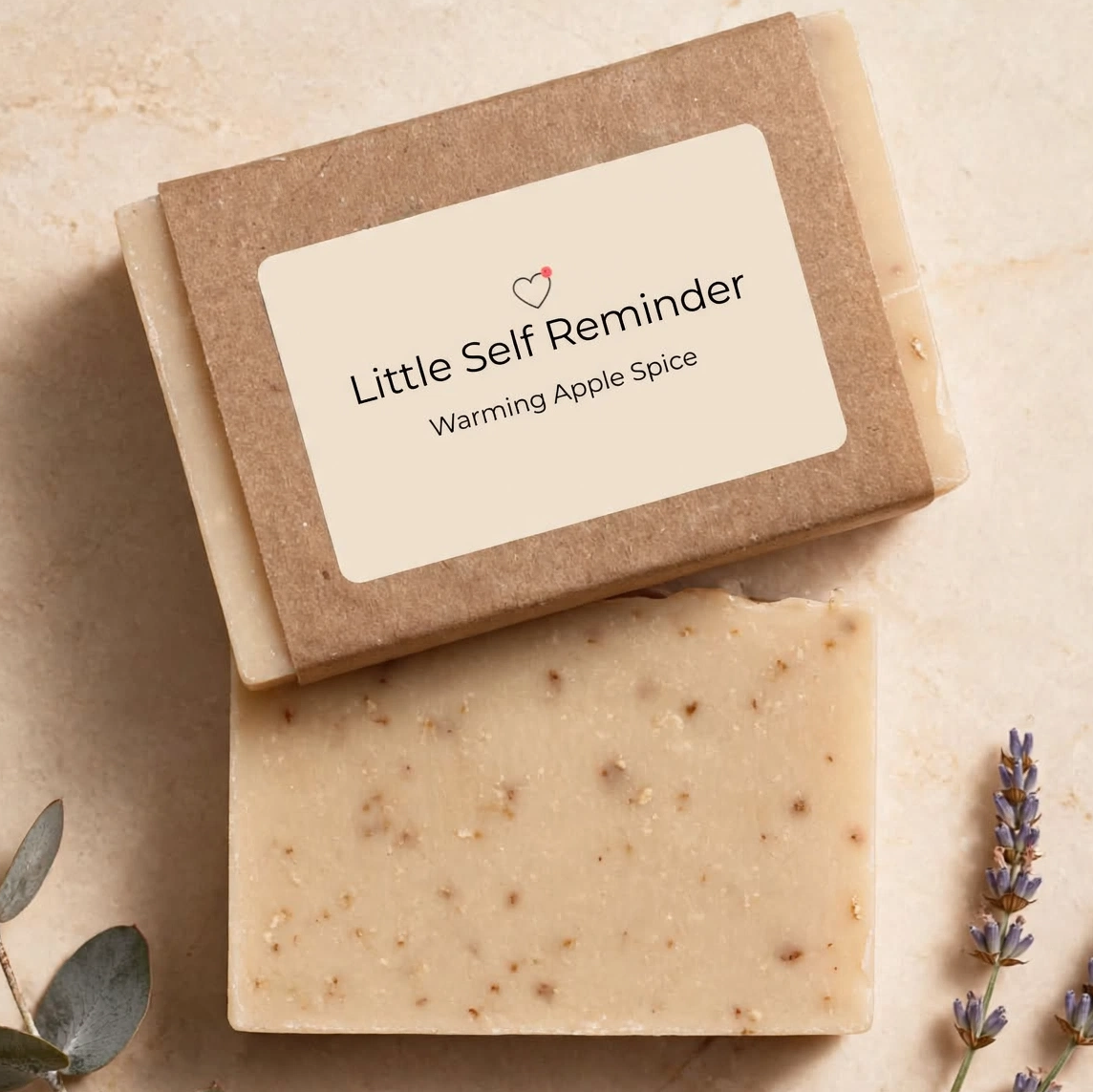 A bar of soap labeled 'Little Self Reminder' and 'Warming Apple Spice' stacked on top of its Kraft paper box.